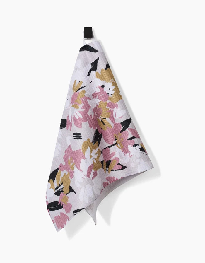 The Toboggan Avenue Wilderness Floral Bar Towel, featuring a pink, white, mustard yellow, and black floral design, hangs on a black loop against a white background. Crafted from durable Geoweave fabric.