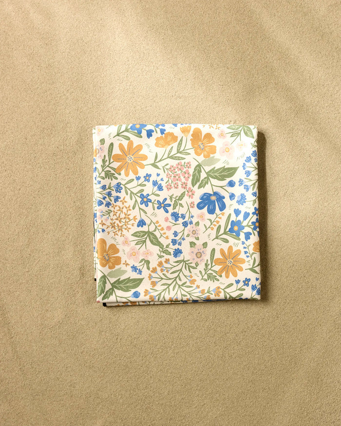 The Danelys Sidron "Wildflower Vine Blush" is a folded square beach blanket with a colorful floral pattern, made from quick-drying recycled materials, shown on a textured beige surface.