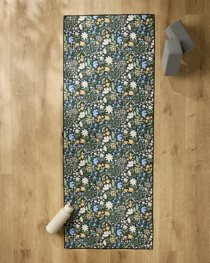 The Danelys Sidron Wildflower Vine Blush yoga mat, featuring white, blue, and yellow flowers, rests on a light wooden floor with a non slip towel, a white water bottle at one end, and two gray yoga blocks nearby.