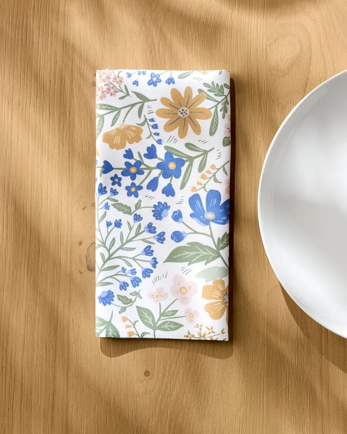 The Danelys Sidron Wildflower Vine Blush napkin set, featuring vibrant florals on Geosuede fabric, is neatly folded on a wooden table beside a white plate. Sunlight highlights the elegant, recycled design.