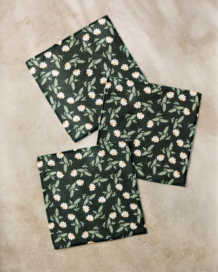 Three Wildflower Vine Onyx cloth napkins by Danelys Sidron, made from recycled materials, display a white floral and green leaf design on dark green fabric, neatly arranged on a beige stone surface.