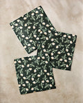 Three Wildflower Vine Onyx cloth napkins by Danelys Sidron, made from recycled materials, display a white floral and green leaf design on dark green fabric, neatly arranged on a beige stone surface.