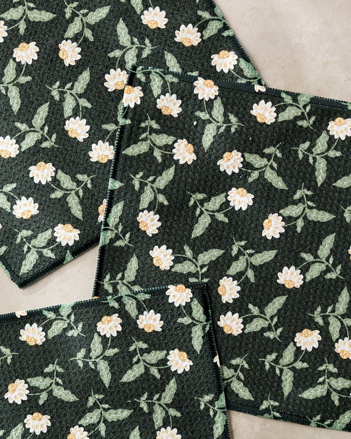Three dark green “Wildflower Vine Onyx” dishcloths by Danelys Sidron, featuring a white and yellow daisy floral pattern, overlap each other on a light beige surface.