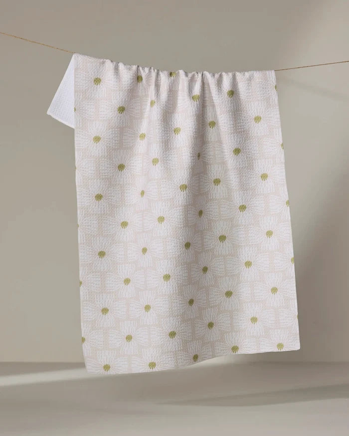 The Ash Cascade Wildflower tea towel, featuring a white textured design with subtle geometric patterns and green floral accents, hangs on a clothesline against a neutral backdrop.