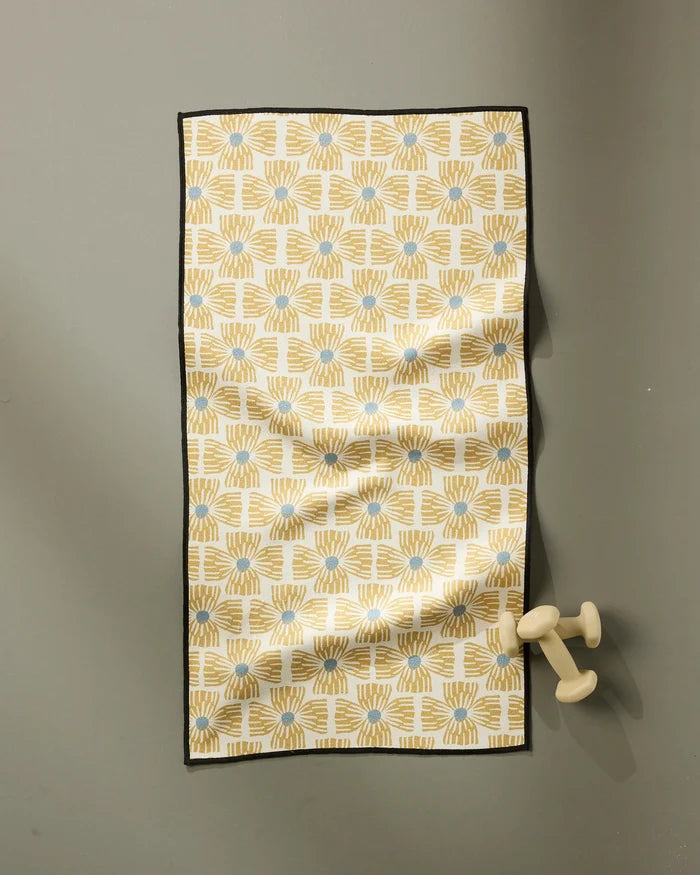 The Ash Cascade Wildflower fitness towel, ultra-absorbent and made from recycled materials, features a yellow and blue geometric pattern with a black border. It hangs on a gray wall with light-colored dumbbells resting on one end.
