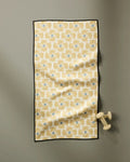 The Ash Cascade Wildflower fitness towel, ultra-absorbent and made from recycled materials, features a yellow and blue geometric pattern with a black border. It hangs on a gray wall with light-colored dumbbells resting on one end.