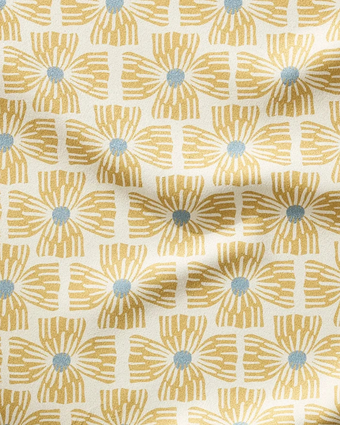 The Ash Cascade "Wildflower" fitness towel showcases yellow geometric floral patterns with blue centers on white. Made from ultra-absorbent recycled materials, its surface displays gentle folds and subtle shadows.