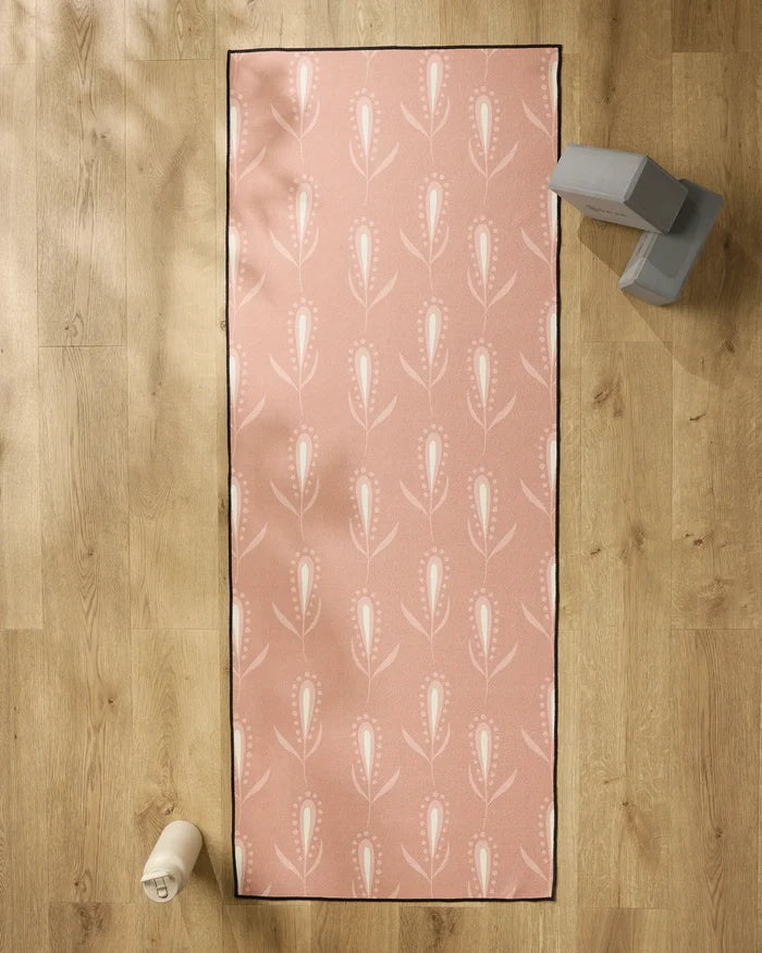 The Amita Nair Wilds pink yoga mat with a white floral pattern is laid out on a wooden floor, topped with a non-slip yoga towel. A water bottle sits to the left, and gray exercise blocks rest at the top right of the mat.