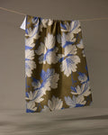 The Geometry Wildshade tea towel, featuring white and blue leaf patterns on a brown background, hangs from a string against a neutral backdrop. Its fabric dries quickly and adds style to any kitchen.