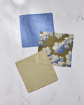 Three folded Geometry Wildshade fabric napkins, made from recycled materials, are displayed on white marble: one blue, one beige, and one brown with a blue and white leaf pattern.