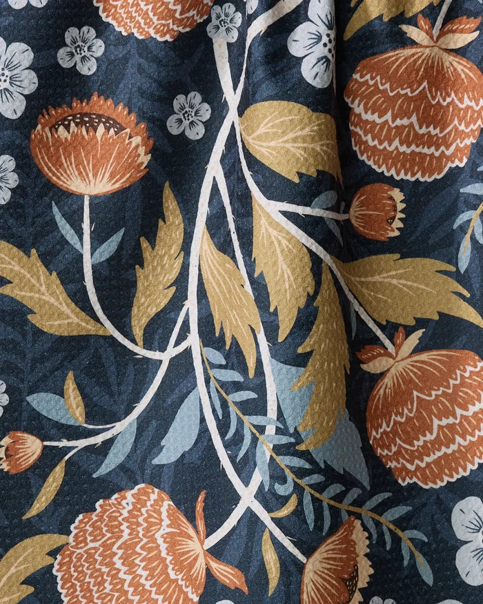 The William tea towel by Danelys Sidron features bold orange and cream flowers, yellow leaves, and white blossoms on dark blue. This vintage-inspired botanical pattern is crafted from quick-drying recycled materials for eco-friendly use.