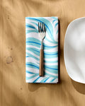 A fork with a wooden handle rests on a Rachel Parker Willow Wave blue and white wavy-patterned reusable table napkin, placed on a light wooden table beside the edge of a white square plate.