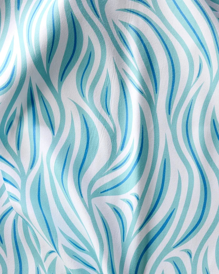 A close-up of Rachel Parker's Willow Wave recycled dinner napkins, featuring an absorbent cloth with a flowing, abstract light blue and turquoise pattern on white, reminiscent of stylized leaves or waves with soft folds and gentle shadows.