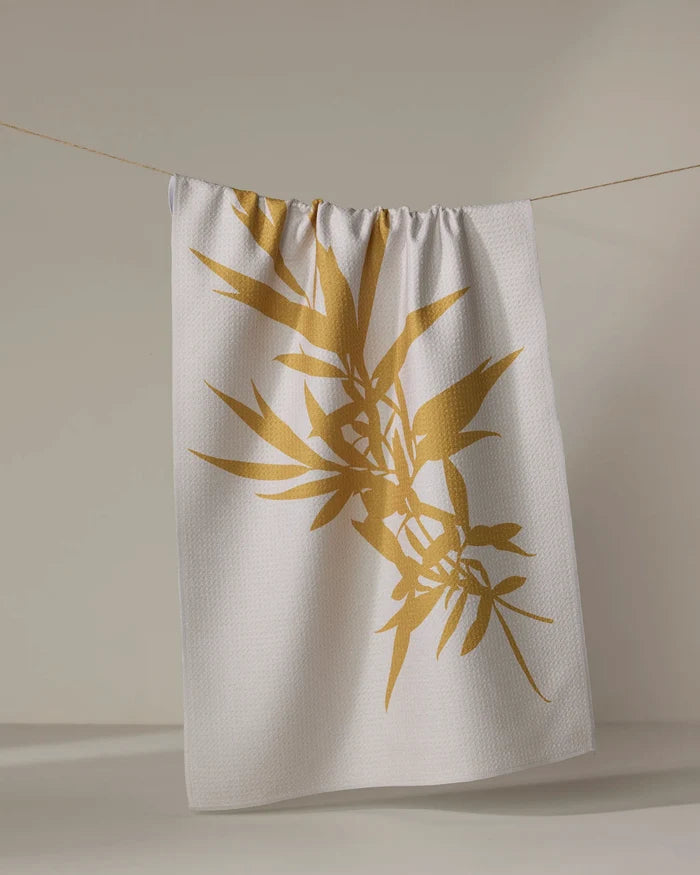 The PrintedPlant Willow towel, featuring a large golden bamboo leaf design, dries quickly and hangs on a clothesline against a neutral background.