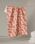 The Wind Fall dish towel by Danelys Sidron, featuring an orange leafy design, hangs against a neutral backdrop. White bowls are stacked in the lower right corner.
