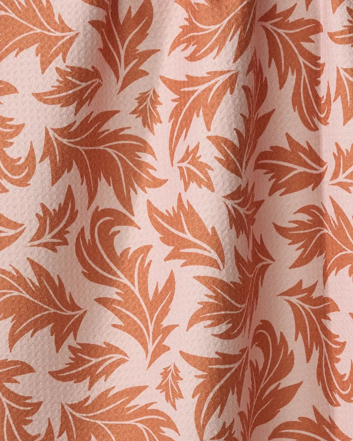 A close-up of the "Wind Fall" dish towel by Danelys Sidron, showing its absorbent textured fabric with a stylized orange leafy pattern on a pale pink background and gentle folds.