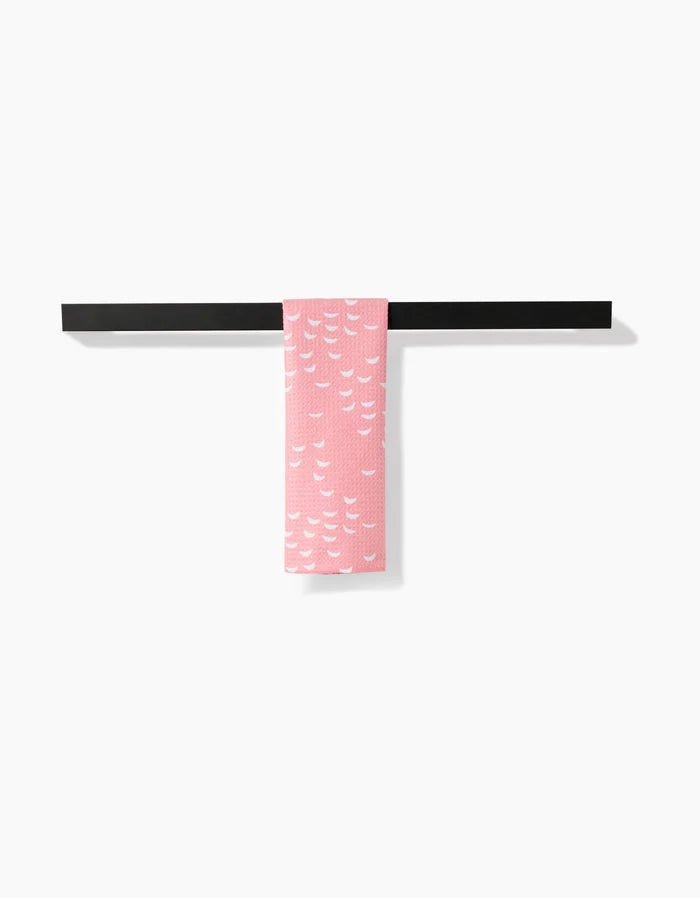 The pink Windswept bar towel by Jessica Swift, made from soft Geoweave fabric and featuring a white wave-like pattern, hangs neatly over a simple black towel bar against a plain white background.