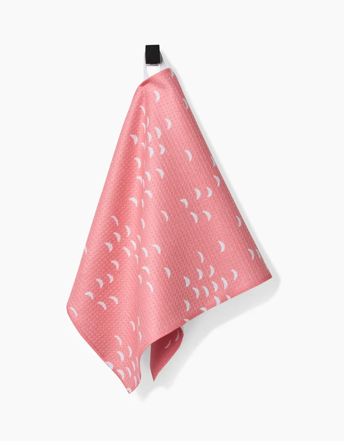 The Windswept bar towel by Jessica Swift, made from recycled materials, features a pink textured fabric with small white crescent shapes and hangs from a black loop against a white background.