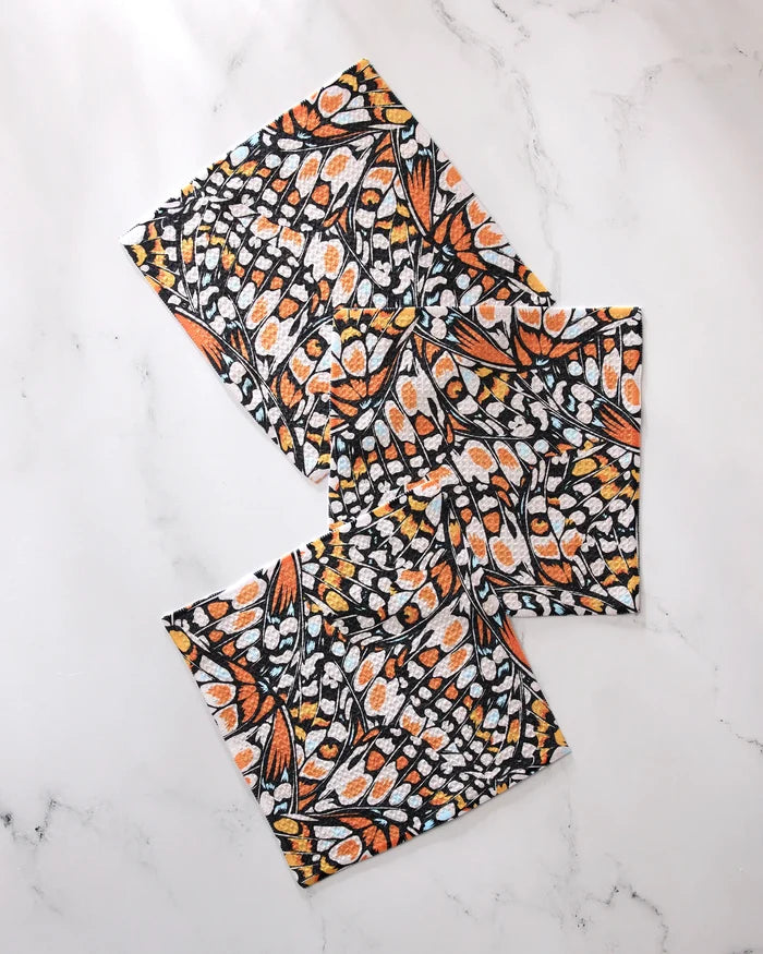 Three "Wings in the Wind" fabric napkins by GEOMETRY, featuring a vibrant monarch butterfly wing pattern in orange, black, yellow, and white recycled material, are arranged in a scattered layout on a white marble surface.
