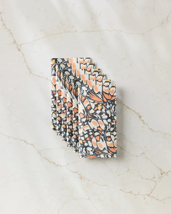 A stack of GEOMETRY’s Wings in the Wind patterned napkins, featuring an abstract butterfly wing design in orange, black, and white, crafted from eco-friendly kitchen towels and displayed on a light marble surface.