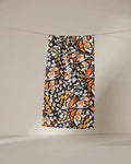 The GEOMETRY Wings in the Wind bar towel, featuring an orange, black, and white butterfly wing pattern, hangs on a thin line against a neutral background.