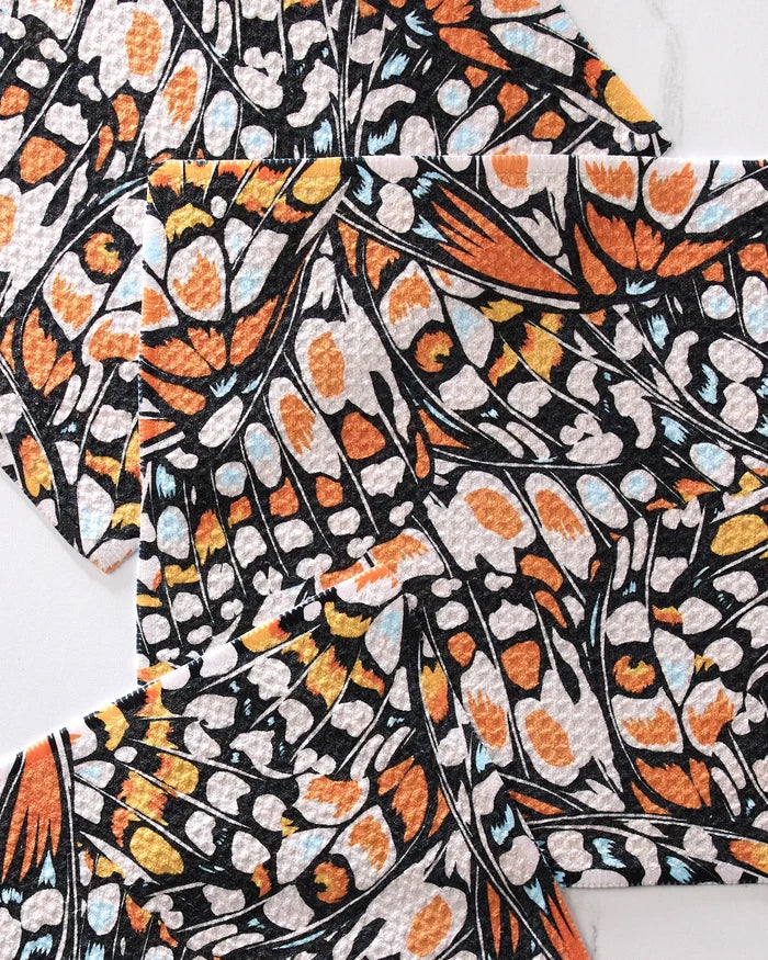 Close-up of overlapping GEOMETRY Wings in the Wind dishcloths, showing a butterfly wing pattern in black, orange, white, and blue on a light background—made from recycled materials.