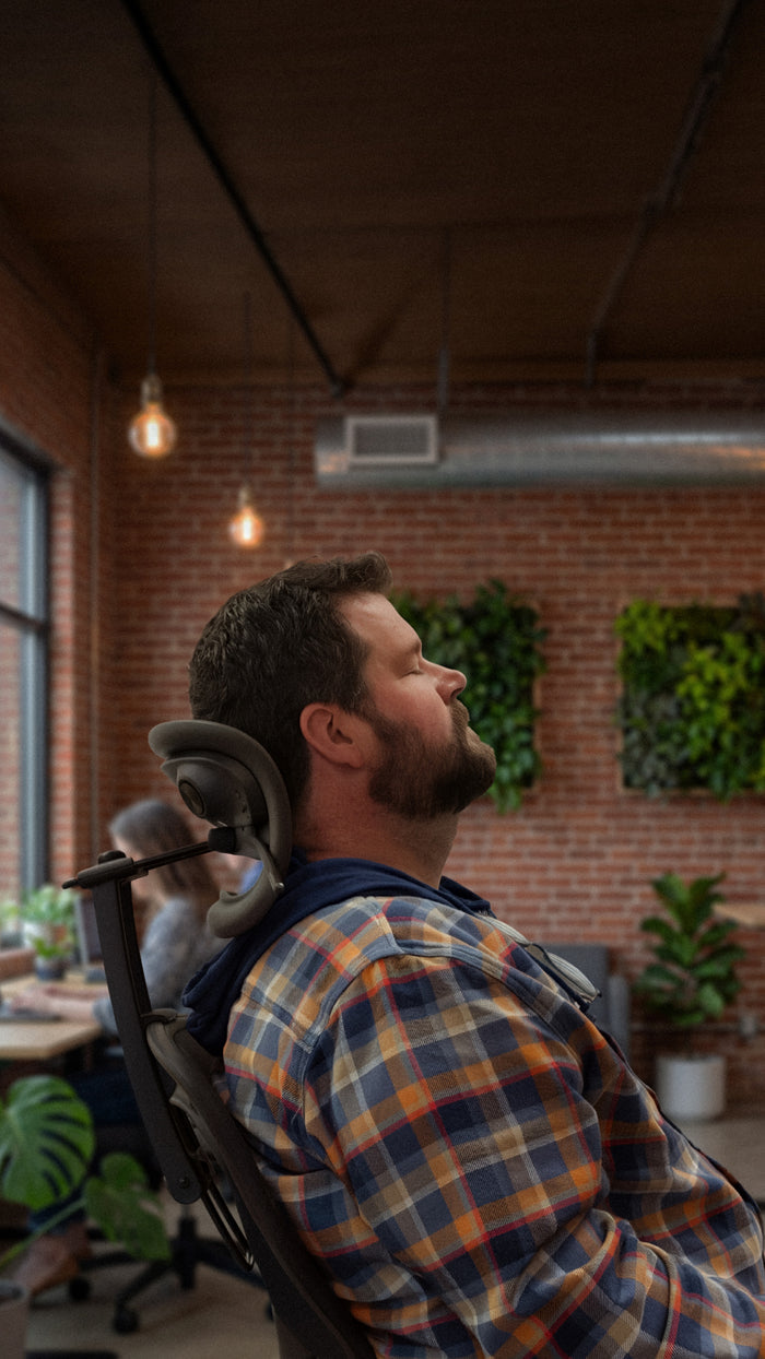 Side view of the Graphite Atlas Aeron Chair Headrest Bluetooth Speaker installed on a Herman Miller Aeron chair and a man relaxing with it.