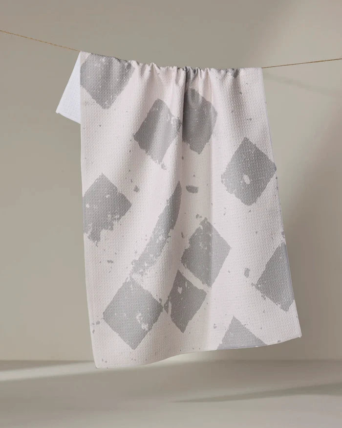 The Geometry Wisp Gray recycled kitchen towel, featuring large irregular gray squares on white, hangs on a clothesline against a neutral background.