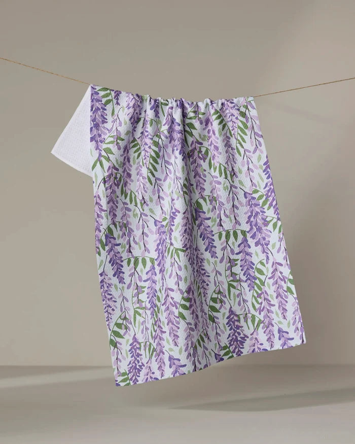 The Rebecca Bobko "Wisteria" kitchen towel, featuring a purple and green floral pattern, hangs on a clothesline against a plain beige background.