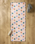 The Claire Iglesias "Wonky Dots" yoga mat, featuring colorful circles, is laid on a wooden floor with a non-slip towel on top. A white water bottle sits at the bottom left, while two gray yoga blocks are stacked in the top right corner.