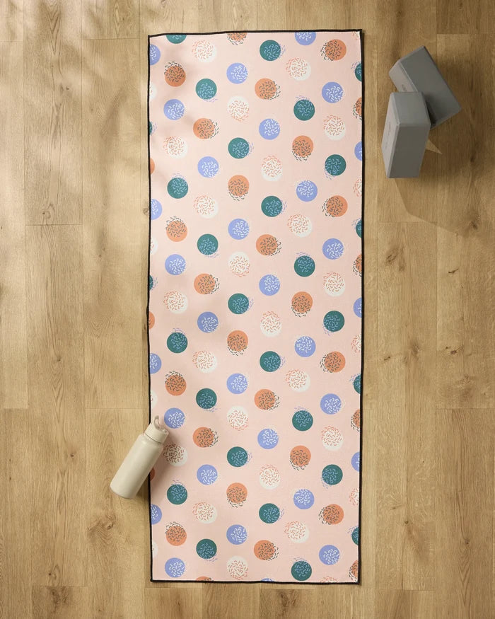 The Claire Iglesias "Wonky Dots" yoga mat, featuring colorful circles, is laid on a wooden floor with a non-slip towel on top. A white water bottle sits at the bottom left, while two gray yoga blocks are stacked in the top right corner.
