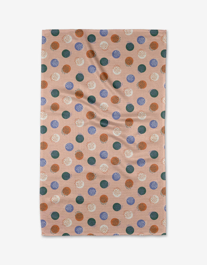 The Claire Iglesias Wonky Dots kitchen towel is beige and rectangular, featuring a pattern of evenly spaced blue, white, orange, and green polka dots with a subtle textured finish and slight creases.