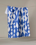 The Claire Iglesias "Wonky Lines" towel, featuring a blue and white wavy abstract design, hangs neatly on a clothesline against a plain background.