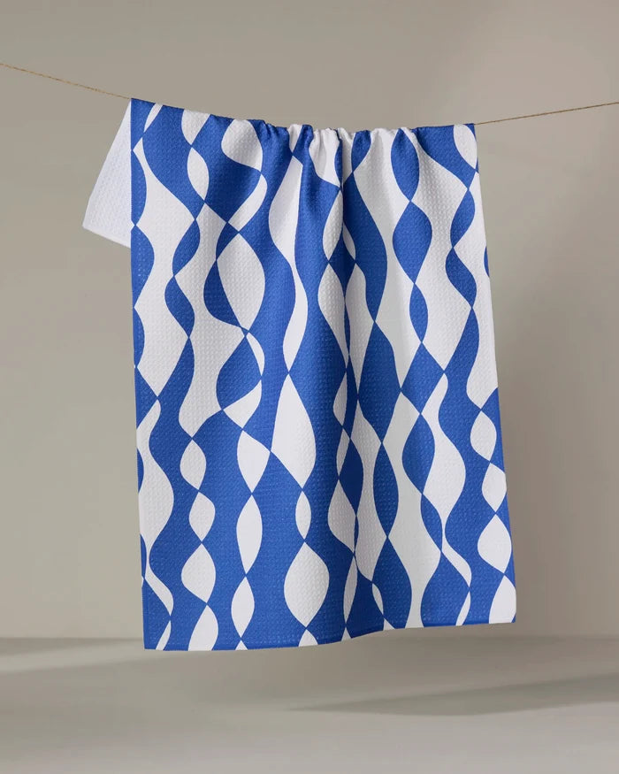 The Claire Iglesias "Wonky Lines" towel, featuring a blue and white wavy abstract design, hangs neatly on a clothesline against a plain background.