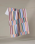 The "Wonky Stripes" kitchen towel by Claire Iglesias, featuring bold red, blue, orange, green, and white geometric stripes, hangs on a clothesline with a plain beige backdrop.