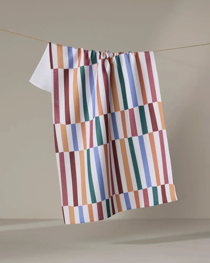 The "Wonky Stripes" kitchen towel by Claire Iglesias, featuring bold red, blue, orange, green, and white geometric stripes, hangs on a clothesline with a plain beige backdrop.