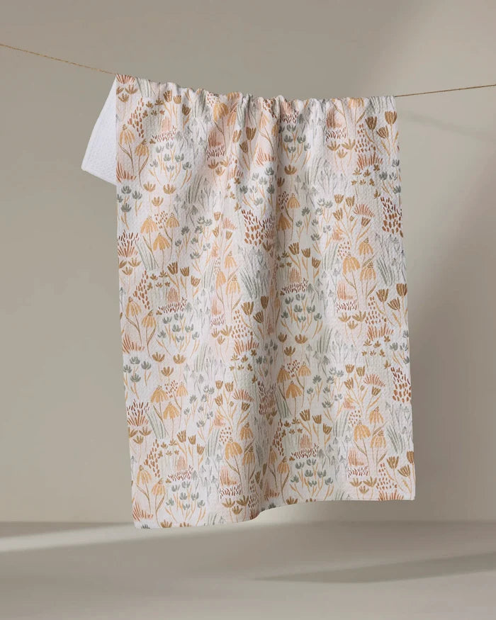 The Woodland Floral tea towel by Hufton Studio, featuring a soft beige, orange, and blue floral pattern, hangs on a clothesline against a neutral background.