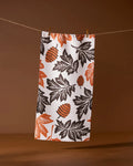The Geometry Woodland Waltz bar towel, made from recycled materials, features an autumn-inspired pattern of orange and black leaves with pinecones and hangs on a clothesline against a brown background.