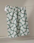 Twig + Spark’s “Woodpile” recycled kitchen towel, featuring a green, white, and gold geometric pattern of overlapping curved lines, hangs neatly on a clothesline against a neutral background.