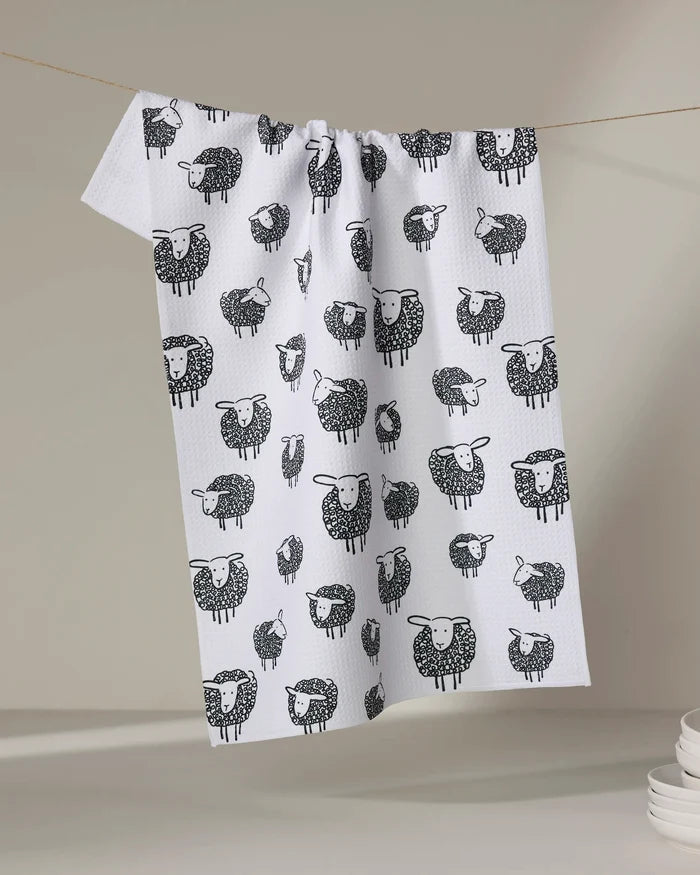 The Kris Ruff Wooly Sheep tea towel, featuring a black and white sheep pattern, hangs on a clothesline against a neutral background with stacked white dishes nearby—showcasing this charming, quick-drying kitchen accessory.