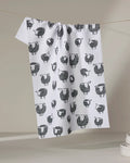 The Kris Ruff Wooly Sheep tea towel, featuring a black and white sheep pattern, hangs on a clothesline against a neutral background with stacked white dishes nearby—showcasing this charming, quick-drying kitchen accessory.