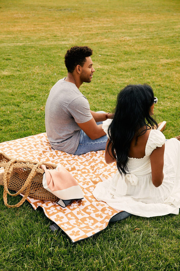 A man and woman picnic on grass atop the GEOMETRY Yellow Block Stripes quick-drying beach blanket. Hes in a gray shirt and jeans; shes in a white dress. Nearby, a woven bag and folded blanket sit beside them, both facing away from the camera.