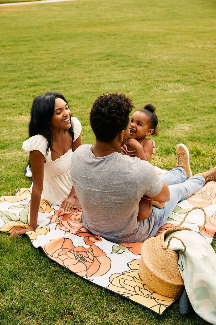 A family of three enjoys a picnic on the grass, sitting atop the GEOMETRY Yellow Checker micro-loop beach blanket. The woman smiles at a man holding a laughing girl, with a straw hat and bag placed nearby.