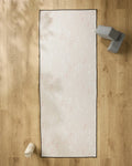 The You Grow Girl yoga mat by Aubrey Fairchild, featuring a white and pink feather pattern, lies on a light wood floor with a slip-resistant towel. Nearby are gray yoga blocks and a white water bottle.