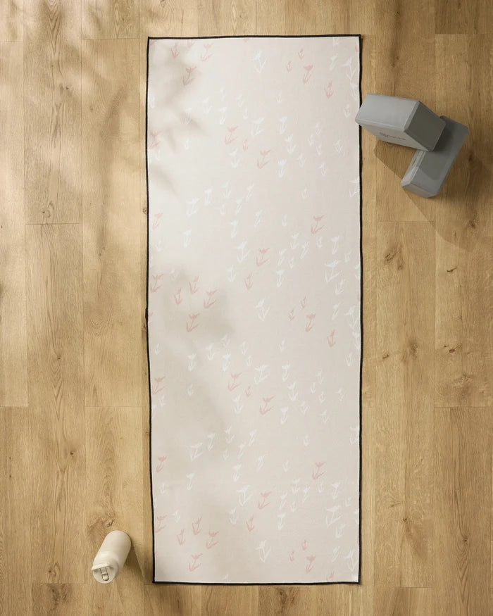 The You Grow Girl yoga mat by Aubrey Fairchild, featuring a white and pink feather pattern, lies on a light wood floor with a slip-resistant towel. Nearby are gray yoga blocks and a white water bottle.