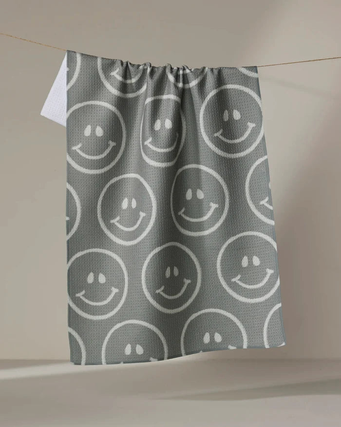 The "You Make Me Happy" towel by Kelsi Fullmer, featuring a gray ultra-absorbent fabric with white smiley faces, hangs on a clothesline against a plain beige background.