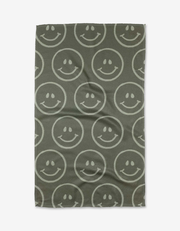 The You Make Me Happy tea towel by Kelsi Fullmer features a gray Geoweave fabric with large, white outlined smiley faces arranged in rows. The rectangular towel has a slightly wrinkled look.
