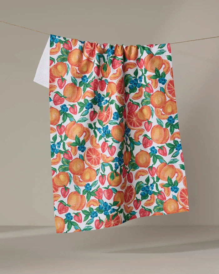 The Zesty Life tea towel by Aubrey Fairchild, made from recycled materials, displays a lively fruit pattern with oranges, strawberries, blueberries, and green leaves as it hangs on a clothesline against a beige background.