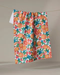 The Zesty Life tea towel by Aubrey Fairchild, made from recycled materials, displays a lively fruit pattern with oranges, strawberries, blueberries, and green leaves as it hangs on a clothesline against a beige background.