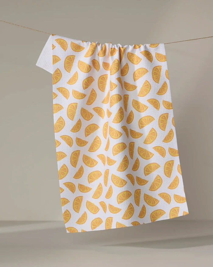 The Aubrey Fairchild Zesty Slice kitchen towel, featuring a vibrant yellow-orange citrus fruit pattern, hangs from a clothesline against a neutral background.